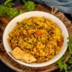 A bowl of Green Pulao garnished with fresh cilantro, served with crispy papad pieces on a wooden platter, with green chilies and a gray napkin in the background.