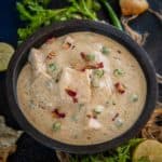 A black bowl filled with creamy Nawabi Paneer curry featuring chunks of paneer and green peas, garnished with herbs and rose petals, surrounded by cilantro, green chili, lemon halves, and torn bread on a dark surface.