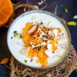 A bowl of orange kheer topped with orange segments and chopped nuts sits on a piece of burlap, with halved oranges and scattered nuts nearby on a dark surface.