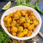A bowl of Aloo Ke Gutke garnished with herbs, surrounded by fresh cilantro, green chili, and lime wedges on a dark surface.