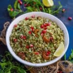 A bowl of Green Poha, garnished with pomegranate seeds, cashews, and fresh herbs, with lemon wedges on the side, placed on a dark surface with scattered greens and herbs around.