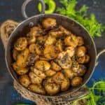 A rustic metal bowl filled with spiced Jakhiya Aloo garnished with herbs, placed on burlap, surrounded by fresh green chilies and coriander on a dark surface.