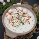 A bowl of creamy makhana raita garnished with whole makhana, chopped fresh herbs, and a sprinkle of red chili powder, set on a rustic surface with loose herbs and makhana around.