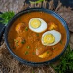 A bowl of Muttai Kulambu, featuring two halved boiled eggs atop a rich, reddish-brown gravy, garnished with herbs and served with flatbread and green chilies on a rustic surface.
