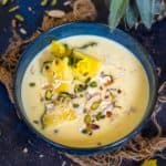 A blue bowl filled with creamy yellow Pineapple Basundi, garnished with pineapple slices, chopped nuts, and shredded coconut, sits on a rustic surface with a pineapple crown nearby.