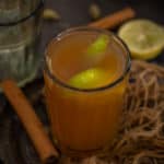A glass of Sulaimani Chai with a lemon wedge inside, placed on a dark surface with a cinnamon stick, cardamom pods, and a cut lemon nearby. An empty glass and textured fabric are in the background.