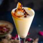 A tall glass of creamy Thandai Frappe, garnished with whipped cream, saffron strands, and dried rose petals, set on a textured mat with scattered rose petals and a small bowl in the background.