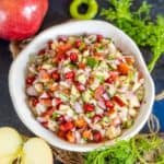 A vibrant bowl of Apple Kachumber featuring chopped apples, onions, tomatoes, cilantro, pomegranate seeds, and green chili, surrounded by whole and sliced apples, green chili, and leafy herbs on a dark surface.