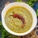 A white bowl filled with Bhang Ki Chutney, garnished with a dried red chili and a drizzle of oil, surrounded by fresh herbs and chili peppers on a dark surface.