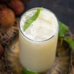 A glass of lychee juice garnished with a fresh mint leaf, sits on a decorative plate surrounded by whole lychees and green leaves.