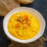 A white bowl filled with creamy yellow Mango Matho, garnished with chopped mango and nuts, placed on a rustic cloth, with crispy puris in the background.