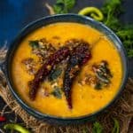 A bowl of Masoor Ki Khatti Dal garnished with dried red chilies, fresh curry leaves, and spices, placed on a textured cloth with green chilies and cilantro around it.