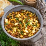 A bowl of spicy Boiled Egg Bhurji garnished with fresh cilantro, served with green chilies, bread, and a glass of tea on the side. The food is arranged on a rustic tray with fresh herbs.
