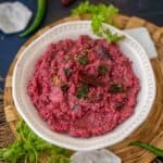 A white bowl filled with vibrant pink Beetroot Curd Rice, garnished with curry leaves and set on a woven mat, with fresh coriander and green chilies nearby.