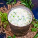 A glass of Kathiyawadi Tadka Chaas garnished with chopped green herbs, placed on a piece of burlap with fresh curry leaves and green chilies in the background.