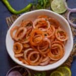 A bowl of spiced Laccha Onion rings garnished with herbs, surrounded by sliced onions, lemon, green chili, and fresh coriander on a dark textured surface.