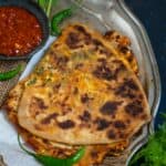 Golden-brown Schezwan Paneer Paratha folded in half is served on a metal tray with a small bowl of red chutney, green chilies, and fresh cilantro garnish.