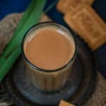 A glass of Lemongrass Chai sits on a saucer atop a burlap cloth, surrounded by rectangular biscuits and a green leaf, set against a dark blue background.