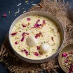 A bowl of Custard Rasgulla, an Indian dessert with cheese balls soaked in creamy, saffron milk and garnished with dried rose petals and chopped nuts, placed on burlap with a small bowl of garnishes beside it.