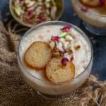 A glass bowl of gulab jamun rabdi parfait topped with sliced gulab jamun, chopped pistachios, slivered almonds, and dried rose petals served beside a small bowl of mixed nuts.