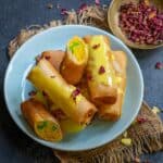 A plate of golden-brown Motichoor Rolls topped with yellow sauce and rose petals, with one roll cut open to reveal a motichoor filling. A bowl of dried rose petals sits in the background.