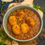 Spicy Anda Matar Masala, served in a light green bowl on a wooden board. Garnished with herbs, green chilies, and naan bread on the side.