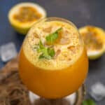 A glass of vibrant Passion Fruit Juice with ice cubes and a mint garnish sits on a rustic surface, surrounded by passion fruit halves and scattered ice cubes in the background.