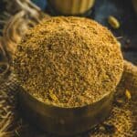 A close-up of a brass bowl filled with a mound of ground brown Tea Masala, placed on a piece of burlap fabric.