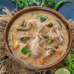 A bowl of creamy Kerala Chicken Stew with vegetables, garnished with herbs, sits on a rustic cloth. Slices of lime, green chili, fresh cilantro, and a red chili pepper are arranged around the bowl on a dark surface.