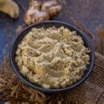A black bowl filled with Ginger Paste sits on a piece of burlap, surrounded by fresh ginger roots on a dark, textured surface.