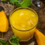 A glass of bright yellow Mango Frooti juice with ice cubes, placed on a burlap cloth, surrounded by fresh mango slices, mint leaves, and extra ice cubes.