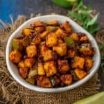 A white bowl filled with spicy, diced mango pickle, Phodnichi Kairi, sits on burlap, surrounded by fresh mango slices, green leaves, and a halved mango in the background.