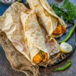 Two rolled Paneer Kathi Rolls filled with paneer, sliced onions, and green chilies rest on parchment paper. Lemon wedges, fresh coriander, and extra green chilies are scattered around for a vibrant garnish.