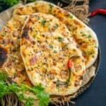 Three pieces of delicious Onion Naan topped with herbs and chilies are arranged on a plate, accompanied by fresh cilantro and a bowl of chopped onion salad in the background.