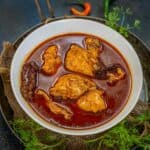 A white bowl filled with pieces of Kashmiri chicken rogan josh in a rich, red curry sauce sits on a rustic tray, garnished with fresh herbs and surrounded by green and red chilies.