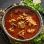 A bowl of spicy red Lamb Rogan Josh with tender pieces of meat, garnished with fresh cilantro, whole green chilies, and lemon slices on the side, served on a rustic dark surface.