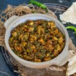 A bowl of Lauki Methi sabji sits on a rustic mat, with green chilies and pieces of roti on the side.