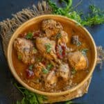 A bowl of spicy Mughlai Chicken Stew garnished with fresh cilantro sits on a rustic cloth, surrounded by red and green chilies and sprigs of cilantro on a dark surface.