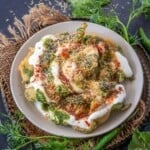A plate of palak patta chaat topped with yogurt, sev, and spices, garnished with fresh cilantro, kept on a rustic mat with green leaves and chilies, all set against a dark background.