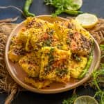 A plate of yellow, spongy Poha Dhokla garnished with herbs and black mustard seeds, surrounded by green chilies, cilantro, and lemon slices on a dark surface.