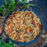 A bowl of Shahi Pulao garnished with cashews, surrounded by fresh green chilies, coriander, and whole spices on a dark rustic surface.