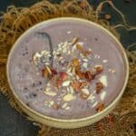 A bowl of creamy Black Rice Kheer topped with chopped nuts and dried rose petals, placed on a piece of burlap fabric over a dark surface. A spoon rests inside the bowl.