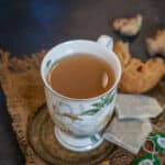 A floral teacup filled with Ginger Green Tea sits on a rustic saucer, surrounded by two tea bags and pieces of biscotti on a burlap mat. Crumbled biscotti and ginger are in the background on a dark surface.