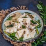 A plate of white, square pieces of Rava Dhokla garnished with mustard seeds, curry leaves, and green chilies, placed on a rustic mat with fresh curry leaves beside it.