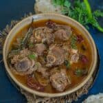 A bowl of spicy Mughlai mutton curry garnished with herbs, surrounded by green chilies, fresh coriander, and pieces of flatbread on a dark background.