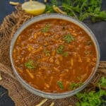 A bowl of spicy Shev Bhaji, garnished with fresh cilantro and surrounded by sev, green chili, lemon wedge, and fresh coriander on a rustic mat.