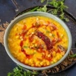 A bowl of yellow boondi kadhi garnished with red chili oil, dried red chili, and fresh coriander sits on a rustic tray, surrounded by green chilies and sprigs of coriander.