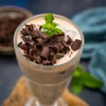 A tall glass of Chocolate Lassi topped with chocolate shavings and a sprig of mint, set on a rustic mat with a blurred background featuring a blue cloth and mint leaves.