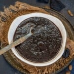 A white bowl filled with thick, dark Kokum Chutney sits on burlap fabric atop a round metal tray. A vintage spoon rests in the bowl, and dried kokum are scattered in the background.