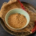 A bowl filled with coarse Raita Masala Powder sits on burlap atop a metal tray. Whole spices and a dried red chili are scattered beside the bowl, all set against a dark background.
